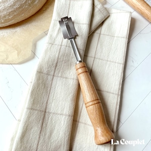 May include: A close-up of a wooden bread lame with a stainless steel blade. The lame is resting on a white and brown checkered cloth. The text "La Couplet" is visible in the bottom right corner of the image.