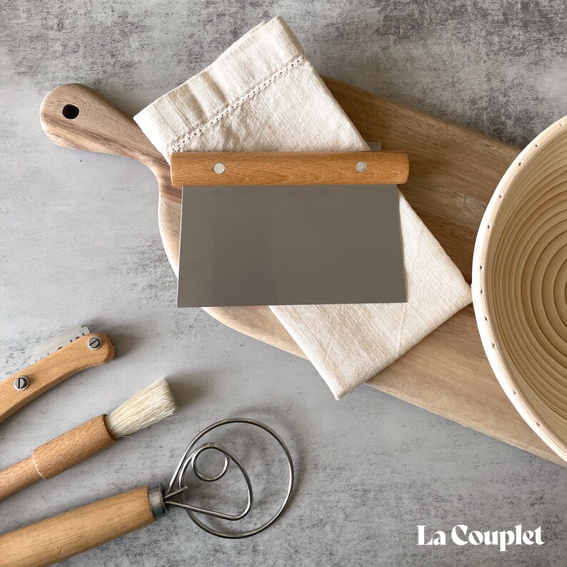 May include: A collection of baking tools on a wooden cutting board. Includes a metal dough scraper with a wooden handle, a linen cloth, a bread lame, a pastry brush, a dough whisk, and a proofing basket. The tools are arranged on a gray surface.