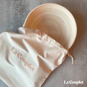 May include: A round, light-colored banneton proofing basket with a spiral design, next to a cream-colored drawstring bag. The bag has the text "La Couplet" printed on it in a soft, peach color. The basket is made of woven material.