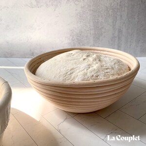 May include: A round, light-colored wooden proofing basket filled with a mound of white dough. The basket has a ribbed design and is set on a white surface. The image is well-lit, highlighting the texture of the dough and the basket's construction.