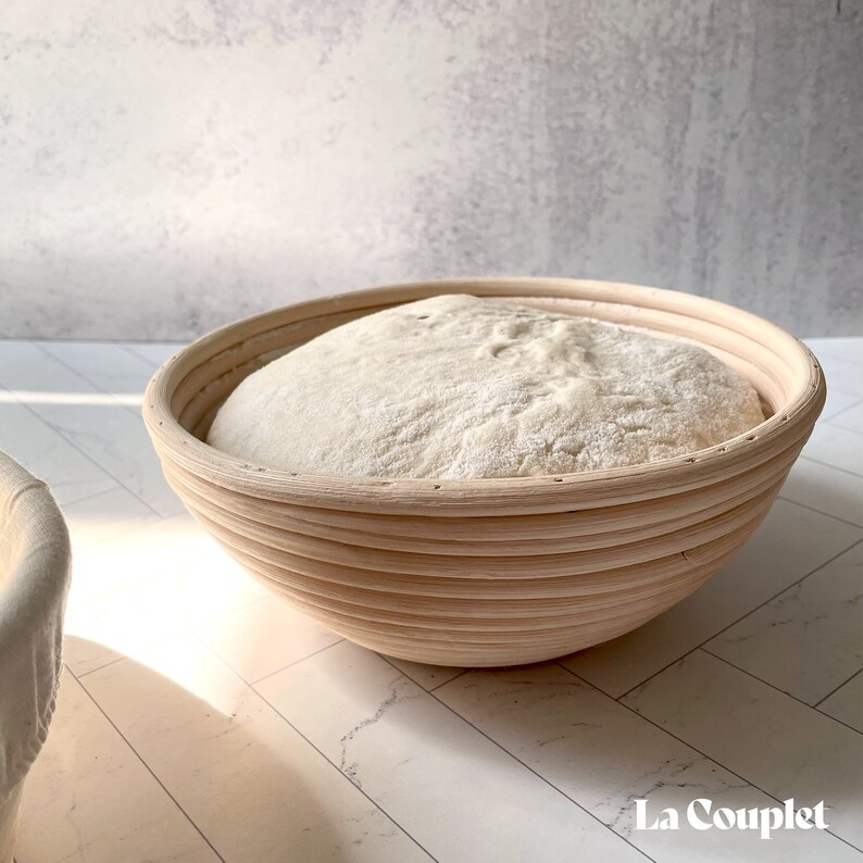May include: A round, light-colored wooden proofing basket filled with a mound of white dough. The basket has a ribbed design and is set on a white surface. The image is well-lit, highlighting the texture of the dough and the basket's construction.