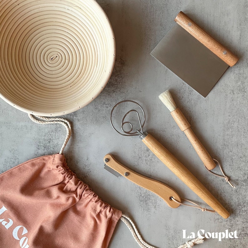 May include: A bread-making kit with a round, coiled proofing basket, a dough scraper, a whisk, a lame, a pastry brush, and a pink drawstring bag. The tools have wooden handles and are arranged on a gray surface. The bag has the text "La Couplet".