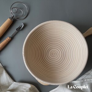 May include: A light-colored wooden banneton proofing basket with a handle, a dough whisk, and a lame. The banneton has a spiral pattern. A white and beige checkered cloth is in the background. The La Couplet logo is in the bottom right corner.
