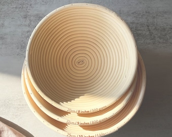 May include: Three round, woven,  wicker bread baskets in different sizes. The baskets are labeled with their dimensions in inches and centimeters, as well as their weight capacity in grams and kilograms. The text reads: 20cm/8 inches/500 grams, 22cm/9 inches/800 grams, 25cm/10 inches/1 kilo. The brand name "La Couplet" is printed below the baskets.