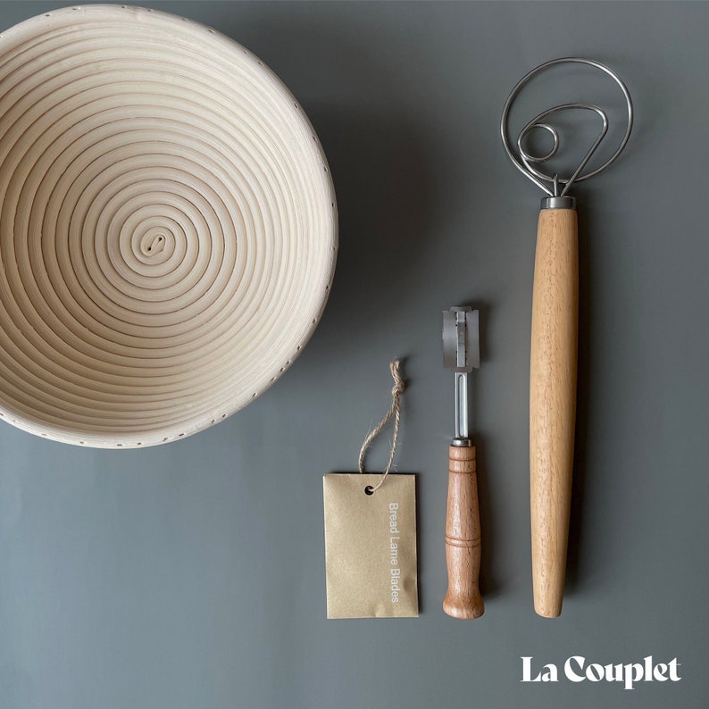 May include: A light-colored wooden banneton proofing basket, a bread lame with a wooden handle, a dough whisk, and a tag that reads "Bread Lame Blades". The items are arranged on a gray surface. The brand name "La Couplet" is at the bottom right.