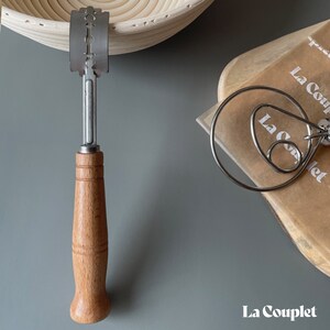 May include: A bread lame with a wooden handle and a stainless steel blade, next to a wire whisk and a proofing basket. The image is on a gray surface. The text "La Couplet" is in the bottom right corner.