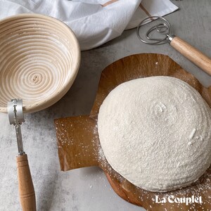 May include: A bread-making setup featuring a round loaf of dough dusted with flour on a wooden board. A proofing basket, a bread lame, and a dough whisk are also visible. The image has a neutral, light-toned background.