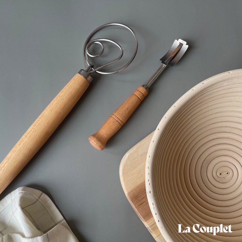 May include: A Danish dough whisk, bread lame, and a proofing basket are displayed on a gray surface. The whisk and lame have wooden handles and stainless steel blades. The proofing basket is made of woven wood. The La Couplet logo is visible.
