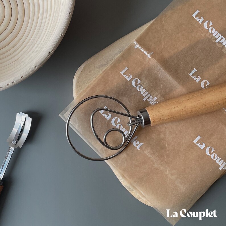 May include: A wooden handled dough whisk and a bread lame with a curved blade rest on a wooden cutting board and parchment paper. The parchment paper is printed with the text "La Couple". A woven bowl is in the background.