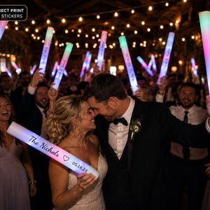 May include: A cylindrical LED foam glow stick featuring a white exterior with the printed text The Nichols, a heart icon, and the date 05.18.24. The stick emits a multi-coloured light, transitioning from blue to pink, and is held by a person.