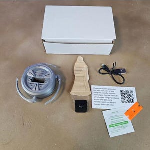 May include: An open white box with a gray circular object, a wooden figure, a black square, a USB cable, and a small orange piece. There are also two small white cards with text and a QR code, and a green packet.