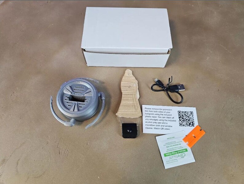 May include: An open white box with a gray circular object, a wooden figure, a black square, a USB cable, and a small orange piece. There are also two small white cards with text and a QR code, and a green packet.