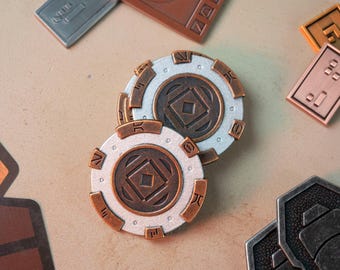 Puede incluir: Dos fichas de color plateado y cobre con un diseño geométrico. Las fichas tienen un fondo blanco con un centro de color cobre y un anillo exterior plateado. Las fichas tienen una forma de diamante en el centro y una serie de círculos concéntricos alrededor del diamante.