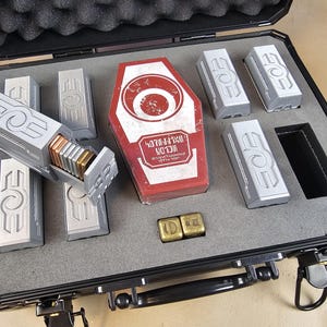 May include: A black case with a gray interior holds several silver rectangular objects with a stylized design. A red and white box with text is also present, along with two gold-colored cubes. The case is open, revealing the contents.
