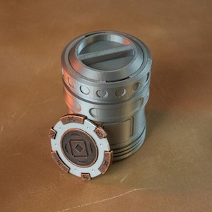 Puede incluir: Un recipiente cilíndrico plateado con tapa a juego y una ficha decorativa circular. La ficha es blanca con detalles en bronce y un diseño geométrico central. El recipiente tiene hendiduras circulares.