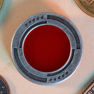 Könnte beinhalten: Ein runder, silberfarbener Metall-Token mit einem roten Zentrum. Der Token hat ein geometrisches Design am Rand.