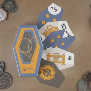 Op de afbeelding: Een set Star Wars-thema speelkaarten en metalen tokens. De kaarten zijn blauw, wit en grijs met verschillende symbolen en nummers. De kaartenbox is blauw en oranje. Verschillende metalen tokens liggen verspreid.