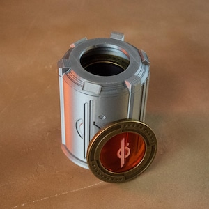 May include: A cylindrical, silver-colored container with a circular opening at the top and a decorative, gold-colored medallion. The medallion has a red center with a white symbol. The container and medallion are on a brown surface.