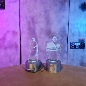 May include: Two clear acrylic light-up figures, one of a robed figure and the other of a man in a suit. Each figure sits on a silver base with a blue light. The background is a textured wall.