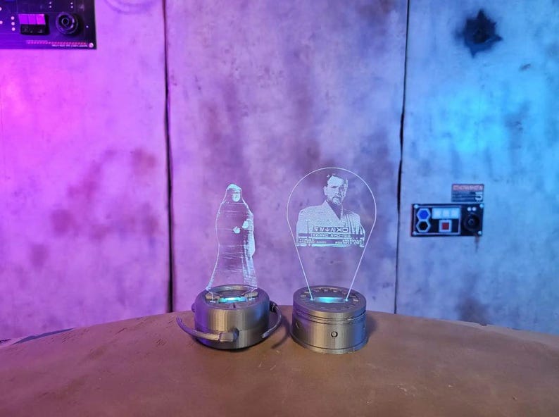 May include: Two clear acrylic light-up figures, one of a robed figure and the other of a man in a suit. Each figure sits on a silver base with a blue light. The background is a textured wall.