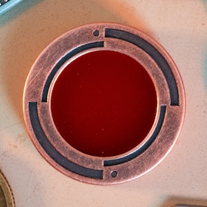 May include: A copper-colored coin with a red center and a black and white design around the edge.