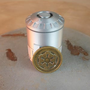 以下が含まれることがあります： 詳細な上部が付いた銀色の円筒形の容器と、円形のエンブレムが付いた金色のコイン。 コインは容器の前に配置されています。 アイテムは石の表面にあります。