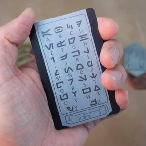 May include: A silver metal card with a black border and a grid of symbols representing the alphabet. The symbols are arranged in rows and columns, with each symbol representing a letter of the alphabet.