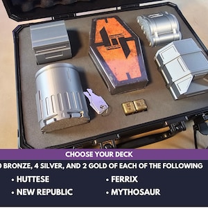 May include: A black case containing various metallic objects, including rectangular and cylindrical components, and two small gold-colored blocks. The case also holds a key and a rectangular box with an orange and black design. Text at the bottom reads "CHOOSE YOUR DECK".