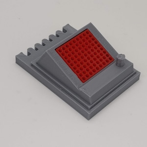 Puede incluir: Un objeto gris impreso en 3D con un patrón de cuadrícula roja en la parte superior. El objeto tiene una pequeña perilla en el lateral.