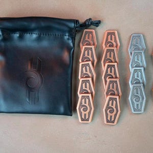 May include: A black drawstring pouch with an embossed design, alongside a collection of metallic tokens. The tokens are in shades of copper, silver, and gold, each with an intricate design. The tokens are shield-shaped.