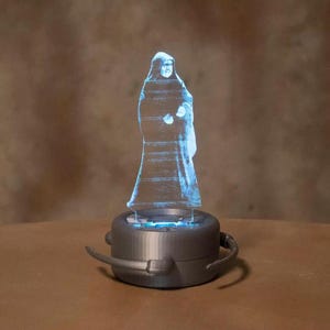 May include: A blue, transparent hologram of a robed figure stands atop a gray, circular base. The base has a dark gray rim and two curved, gray handles. The figure appears to be illuminated from within, creating a glowing effect.