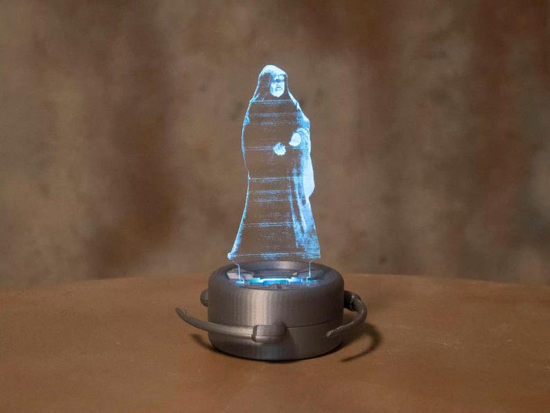 May include: A blue, transparent hologram of a robed figure stands atop a gray, circular base. The base has a dark gray rim and two curved, gray handles. The figure appears to be illuminated from within, creating a glowing effect.