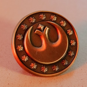 May include: A gold-coloured metal pin featuring a raised design of the Rebel Alliance symbol from Star Wars. The symbol is encircled by a ring of smaller stars.