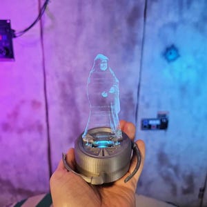 May include: A blue, transparent hologram figure of a person, standing on a grey, circular base with blue lighting. The base has two curved handles. The background is a wall with purple and blue lighting.