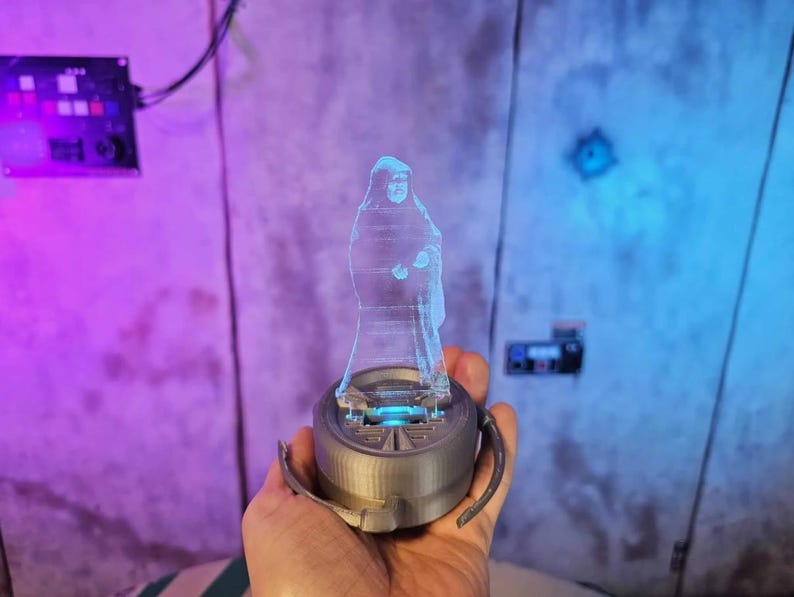 May include: A blue, transparent hologram figure of a person, standing on a gray, circular base with blue lighting. The base has two curved handles. The background is a wall with purple and blue lighting.