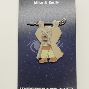 以下が含まれることがあります： スターウォーズのアニメキャラクターが描かれた銀色のエナメルピン。キャラクターは武器を持ち、茶色のローブを着ています。ピンは白の星が散りばめられた青い背景に置かれています。カードの下部には「HYPERSPACE PROPS」と「EO」の文字が印刷されています。