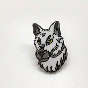 Pode incluir: Um broche de esmalte de cabeça de lobo branco com contornos pretos e olhos amarelos. O lobo está olhando para a direita.