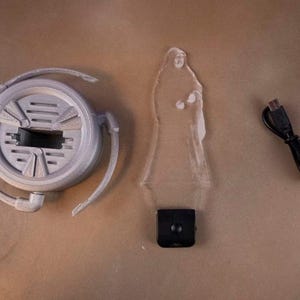 May include: A disassembled gray 3D-printed lamp with a circular base, a clear acrylic figure, and a black USB cable. The lamp base has a central rectangular opening and curved arms. The acrylic figure is a silhouette of a person.