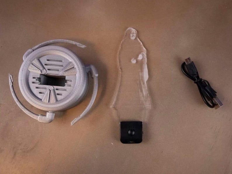 May include: A disassembled gray 3D-printed lamp with a circular base, a clear acrylic figure, and a black USB cable. The lamp base has a central rectangular opening and curved arms. The acrylic figure is a silhouette of a person.