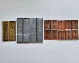 Puede incluir: Un conjunto de 10 fichas de metal con letras en braille. Las fichas están dispuestas en tres filas. La fila superior tiene dos fichas doradas con el número "7" en braille. La fila del medio tiene cuatro fichas plateadas con letras en braille. La fila inferior tiene cuatro fichas de cobre con letras en braille.