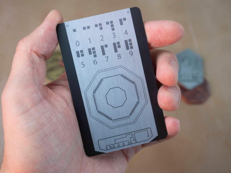 May include: A black and silver card with a grid of numbers 0-9 and a circular design in the center. The card has a black border and a brushed metal finish.