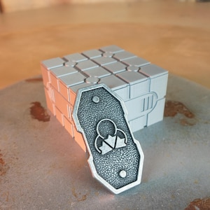 May include: A silver-colored, shield-shaped pendant with a textured surface and a geometric design, resting on a stone surface. Behind it is a silver cube with a grid pattern.