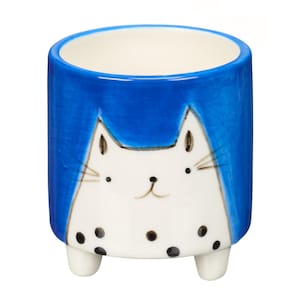 May include: A blue ceramic planter with a white cat face and black polka dots. The planter has three small legs.