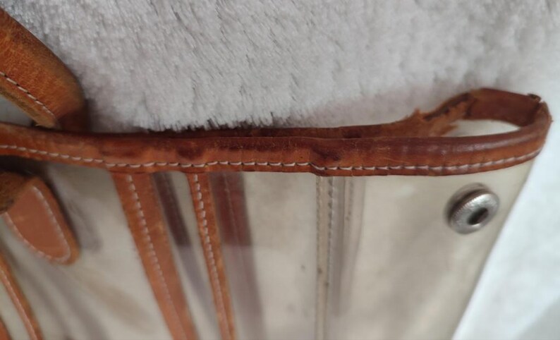 May include: A clear plastic tote bag with a brown leather trim and stitching. The bag has a metal button on the side.