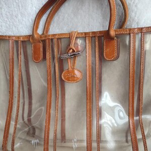 May include: A clear tote bag with brown leather trim and a striped design. The bag has a brown leather handle and a metal clasp.