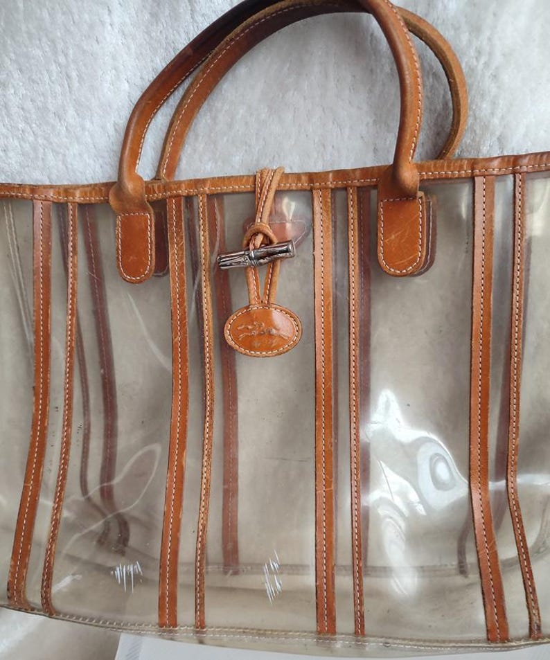 May include: A clear tote bag with brown leather trim and a striped design. The bag has a brown leather handle and a metal clasp.