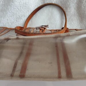 May include: A clear plastic tote bag with brown leather trim and a brown leather handle. The bag has a striped pattern.