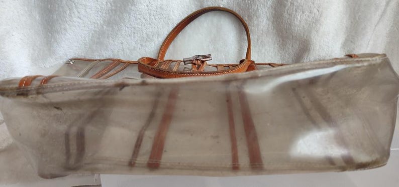May include: A clear plastic tote bag with brown leather trim and a brown leather handle. The bag has a striped pattern.