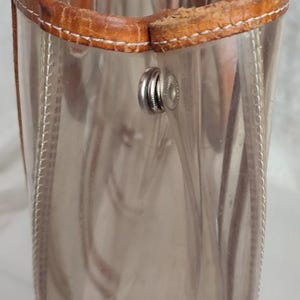 May include: A clear plastic tote bag with brown leather trim and a silver metal clasp. The bag has a zipper closure and a top handle.