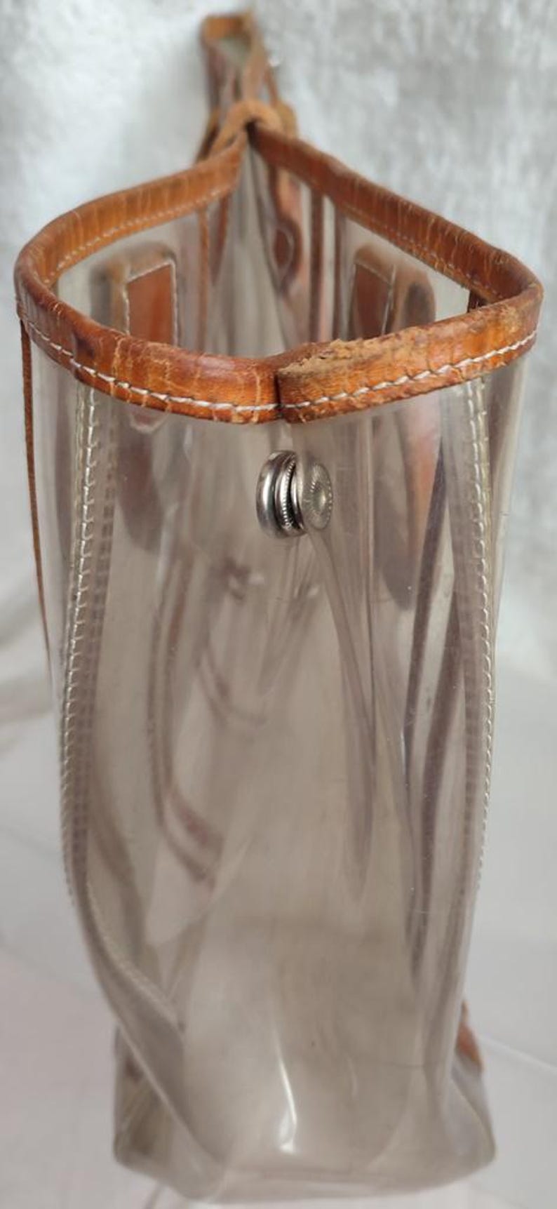 May include: A clear plastic tote bag with brown leather trim and a silver metal clasp. The bag has a zipper closure and a top handle.